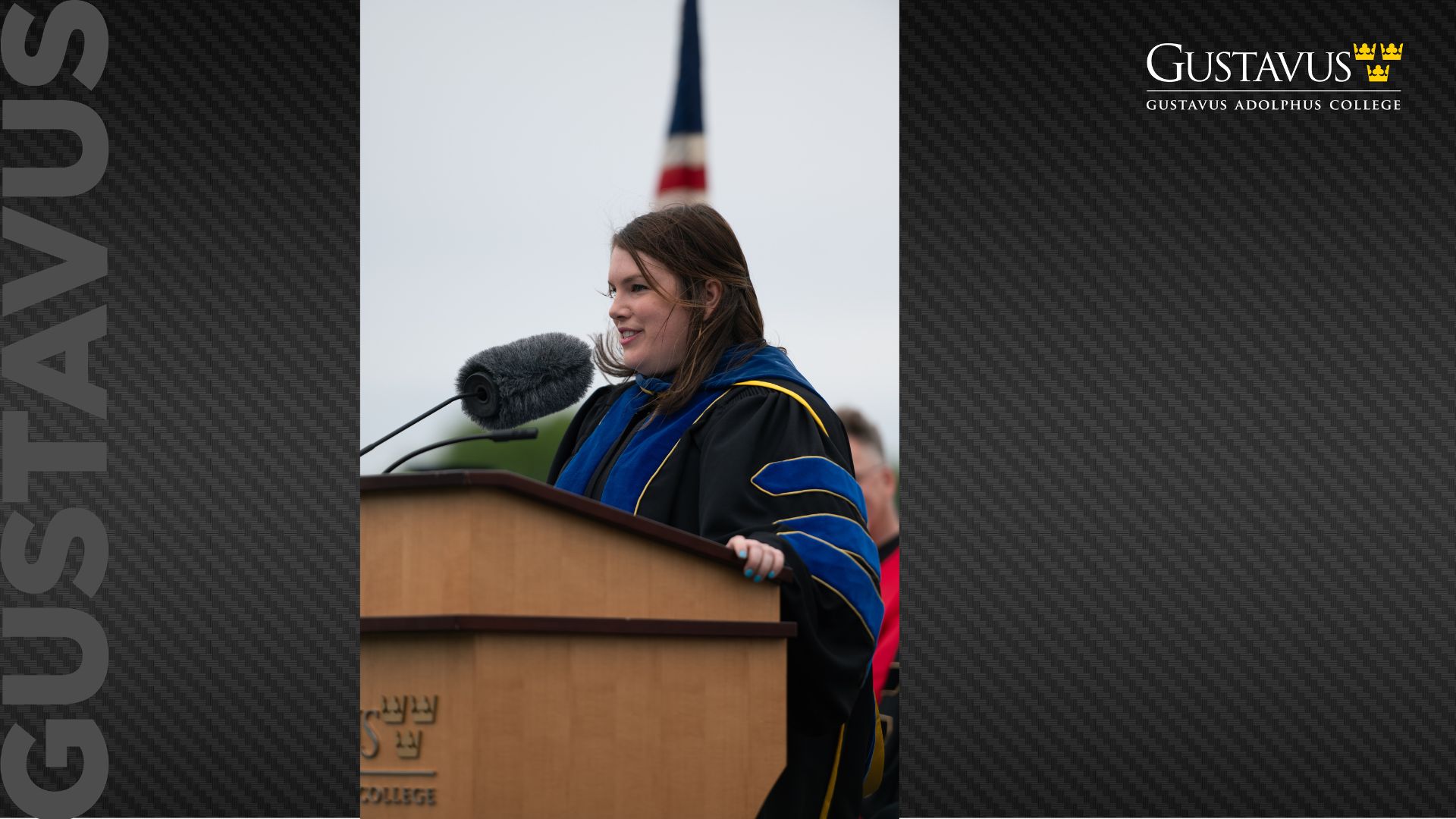 A faculty member in academic regalia speaks at an outdoor podium during a Gustavus Adolphus College ceremony, with an American flag in the background