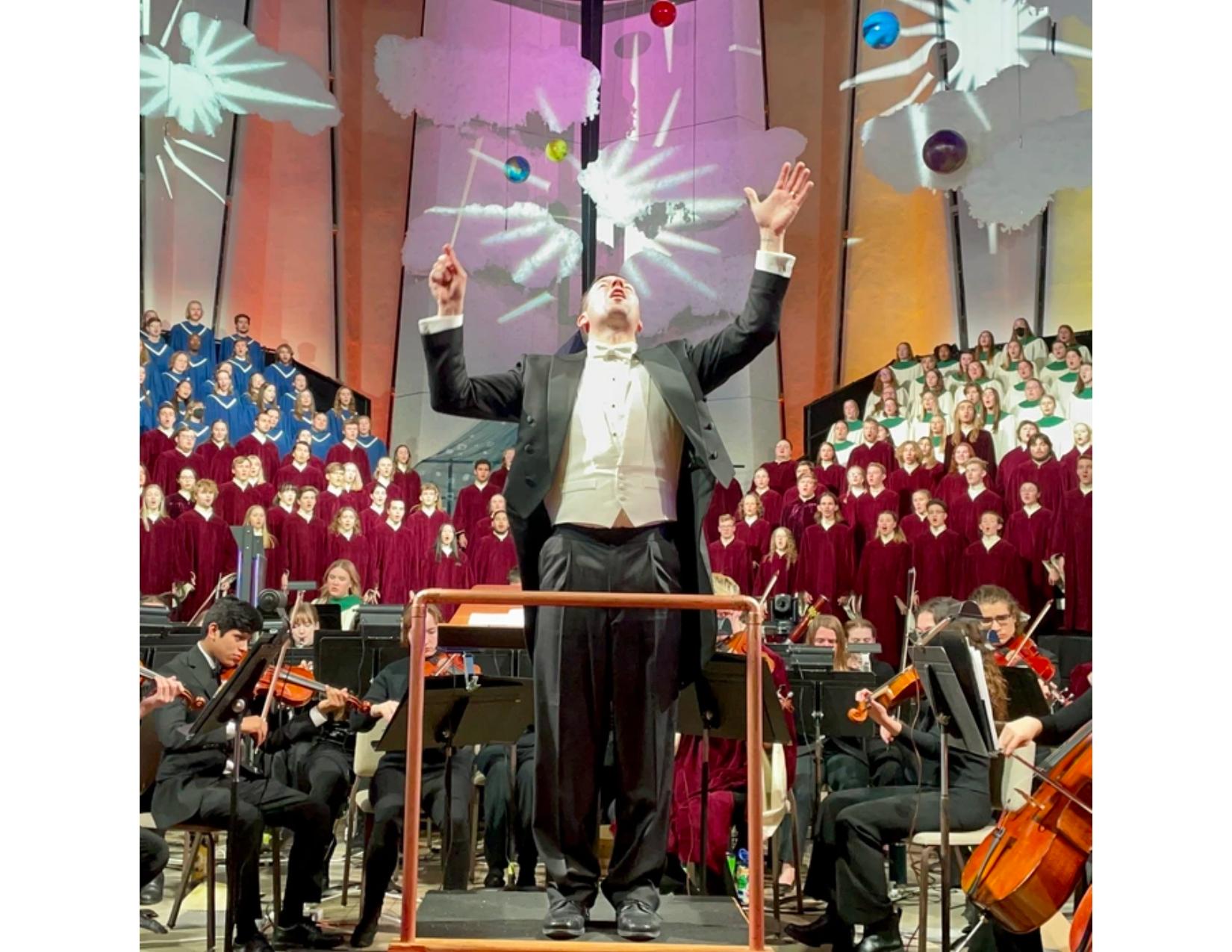 A conductor in formal attire raises his arms mid-performance in front of a large orchestra and choir. The choir is arranged in colored robes by section, blue, burgundy, and white, with dramatic lighting and abstract visuals projected behind them.