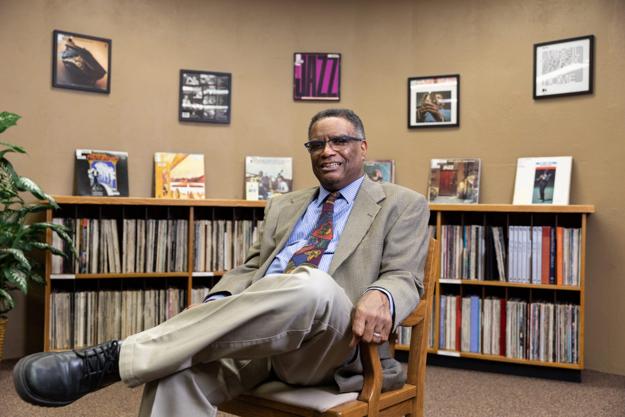 A man in a suit and colorful tie sits smiling with legs crossed in a cozy room lined with records and framed jazz album covers, creating a warm and scholarly atmosphere.
