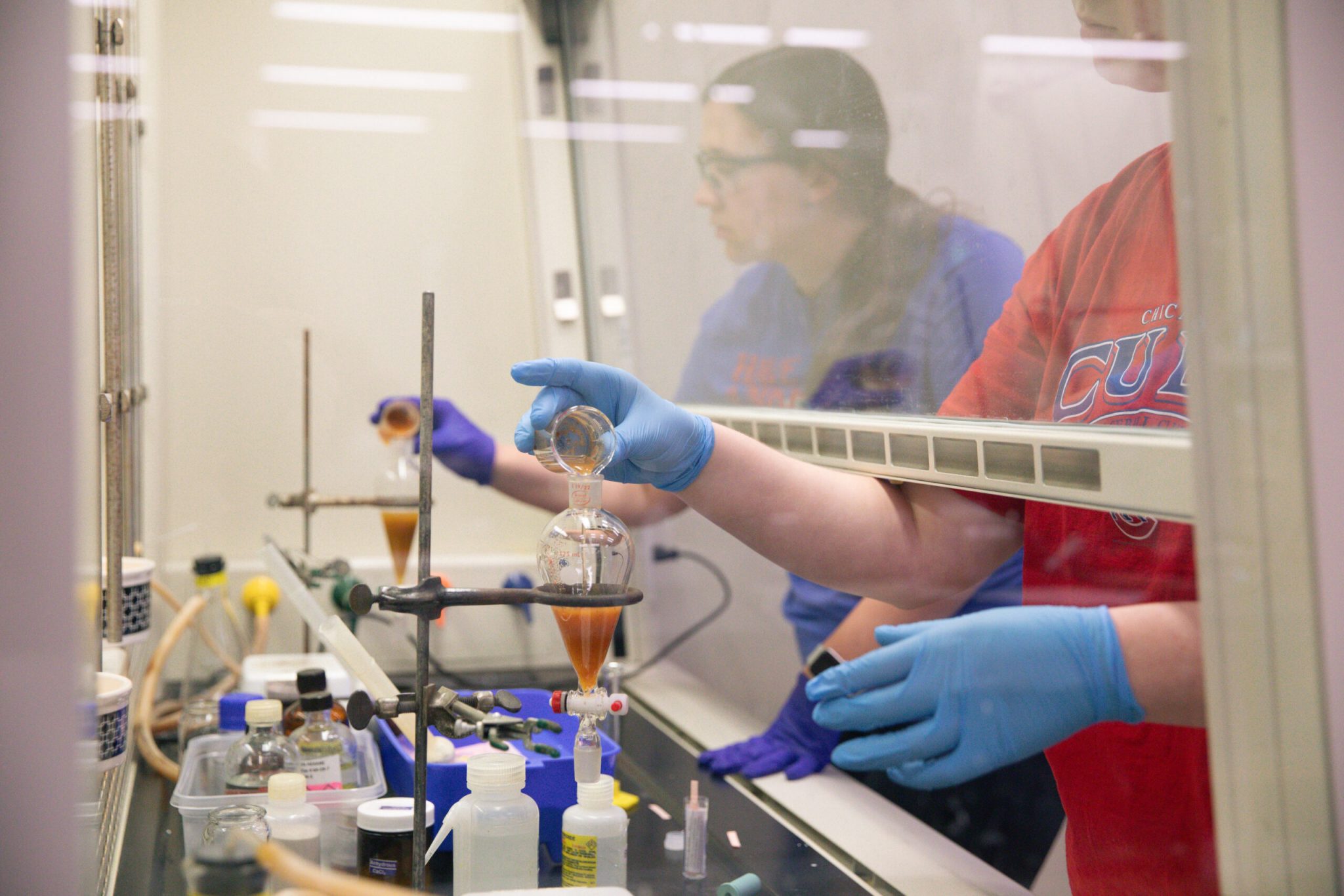 Two students conduct a chemistry experiment in a lab hood, wearing gloves and carefully pouring liquids during a research internship.