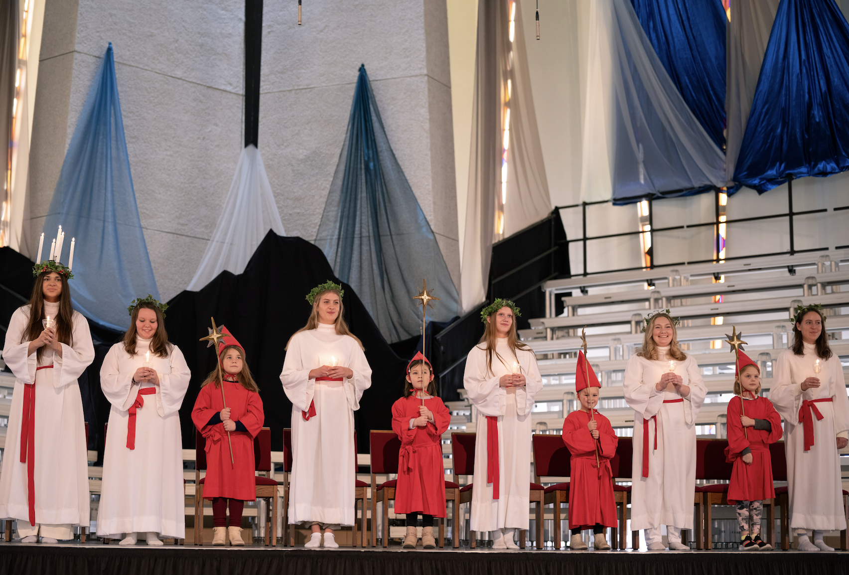 St. Lucia celebration with children and young women in white robes and red sashes, holding candles and star wands on a chapel stage