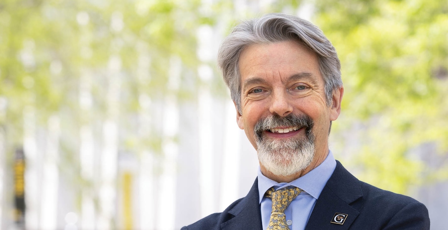 A distinguished gentleman in a suit stands on a college campus in a suit with the G logo of Gustavus Adolphus College pinned to its lapel. 
