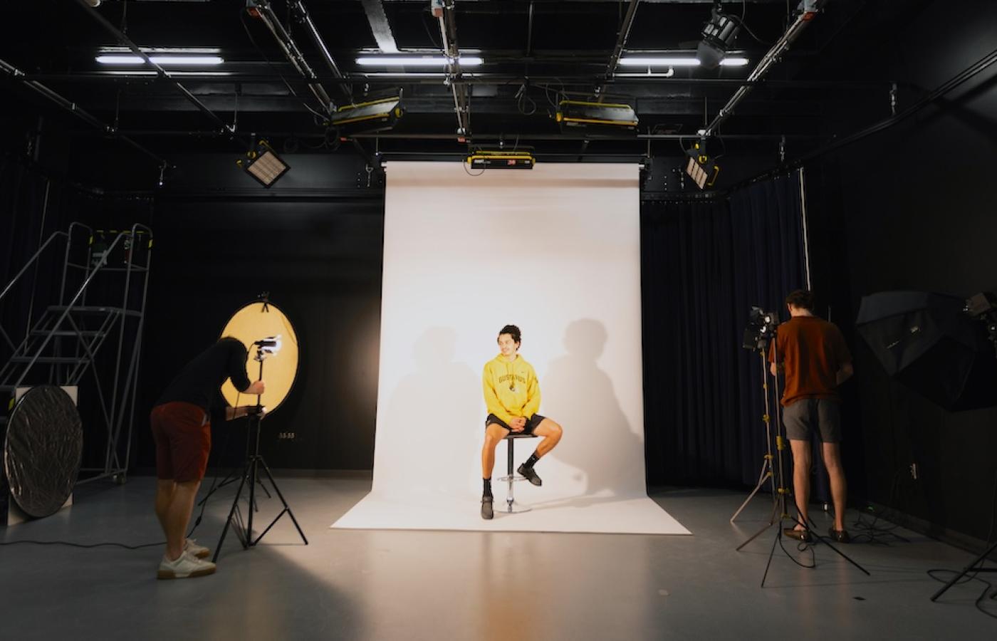 A young man sits on a stool before a white backdrop, in a dark studio. 