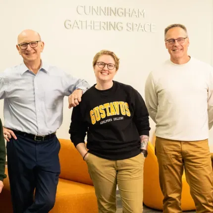Five smiling adults pose together in a brightly lit lounge area labeled 'Cunningham Gathering Space.' They stand in front of orange furniture and a wall-mounted TV, with two of them wearing Gustavus Adolphus College apparel.