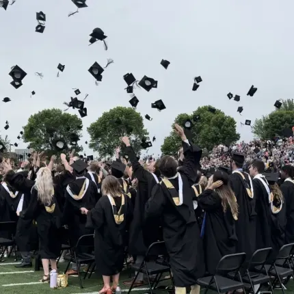 Graduates in black gowns and gold-trimmed stoles throw their caps into the air during the Gustavus Adolphus College 2024 Commencement ceremony, with cheering spectators filling the stadium stands