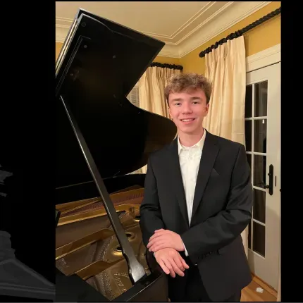 A young man in a suit smiles beside a grand piano in a warmly lit room, with Gustavus Adolphus College branding on the frame