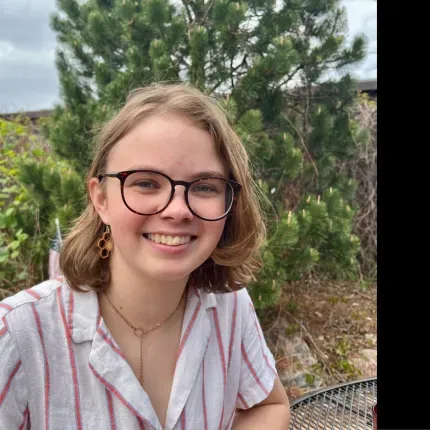 Smiling student seated outdoors with trees in the background, wearing glasses and a striped shirt, with Gustavus Adolphus College branding
