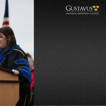 A faculty member in academic regalia speaks at an outdoor podium during a Gustavus Adolphus College ceremony, with an American flag in the background