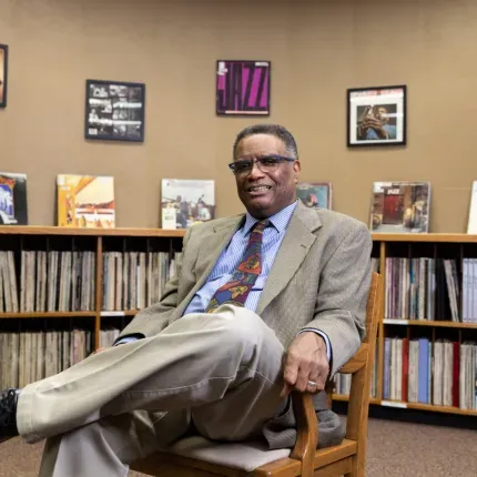 A man in a suit and colorful tie sits smiling with legs crossed in a cozy room lined with records and framed jazz album covers, creating a warm and scholarly atmosphere.