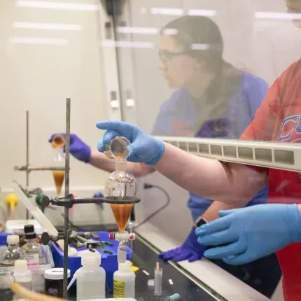 Two students conduct a chemistry experiment in a lab hood, wearing gloves and carefully pouring liquids during a research internship.