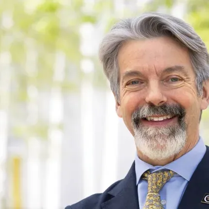A distinguished gentleman in a suit stands on a college campus in a suit with the G logo of Gustavus Adolphus College pinned to its lapel. 