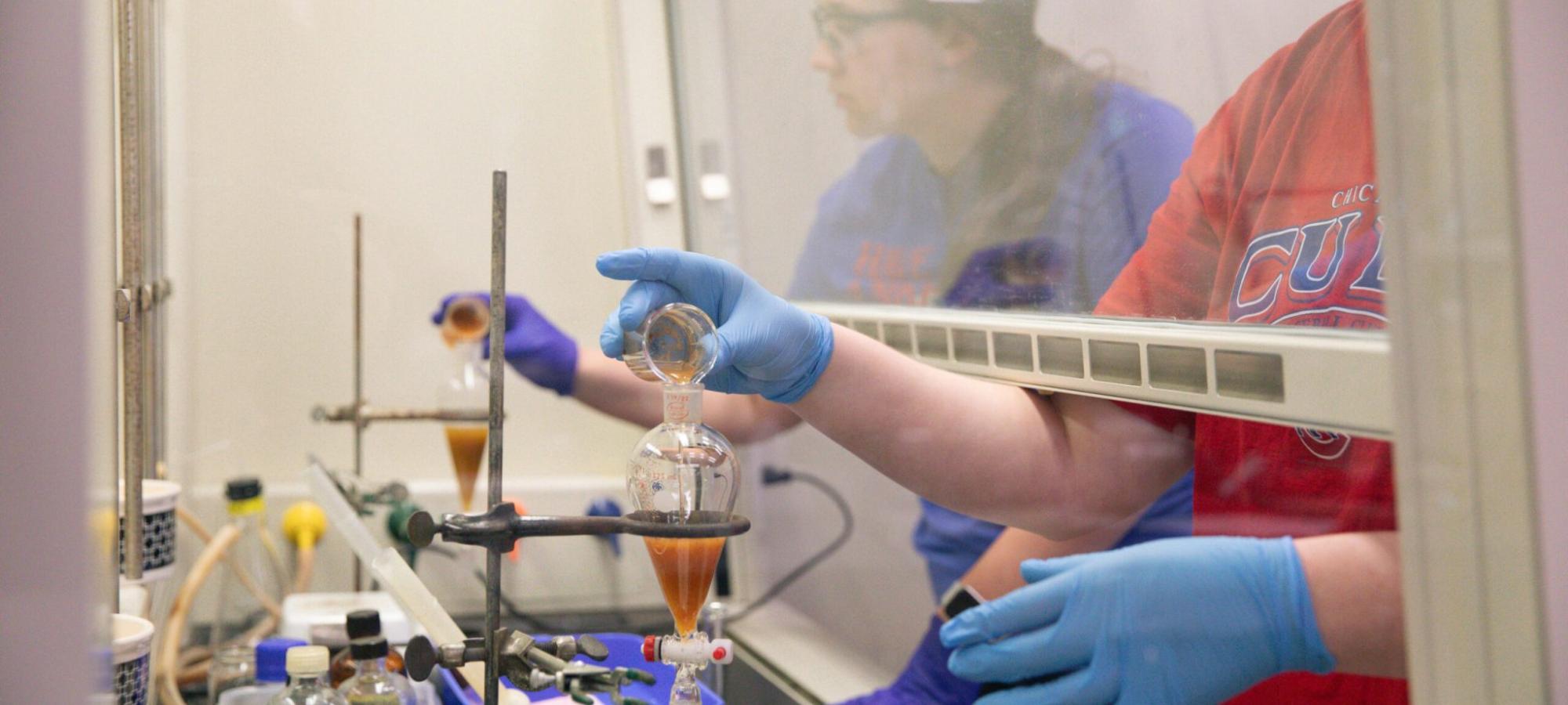 Two students conduct a chemistry experiment in a lab hood, wearing gloves and carefully pouring liquids during a research internship.
