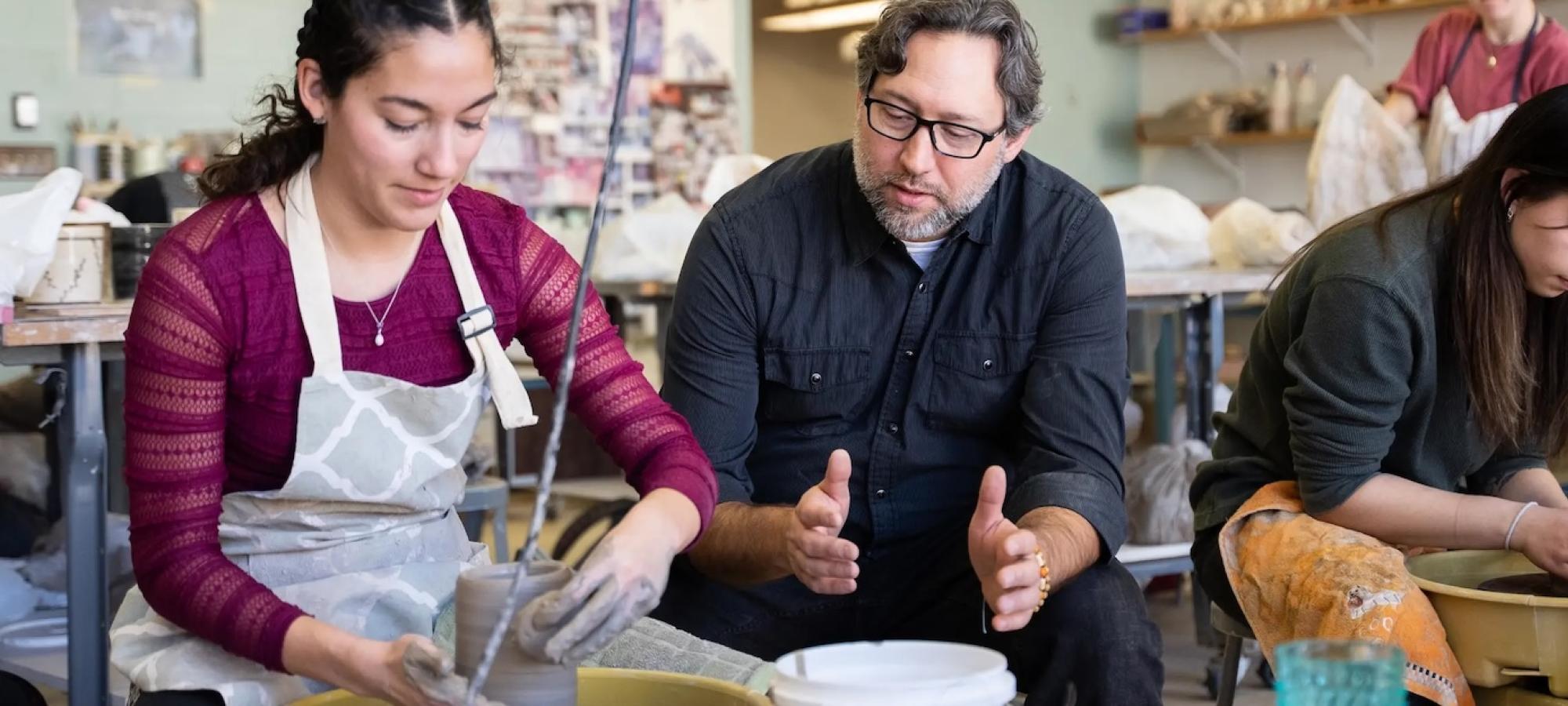 A professor helps a student at a potter wheel. 