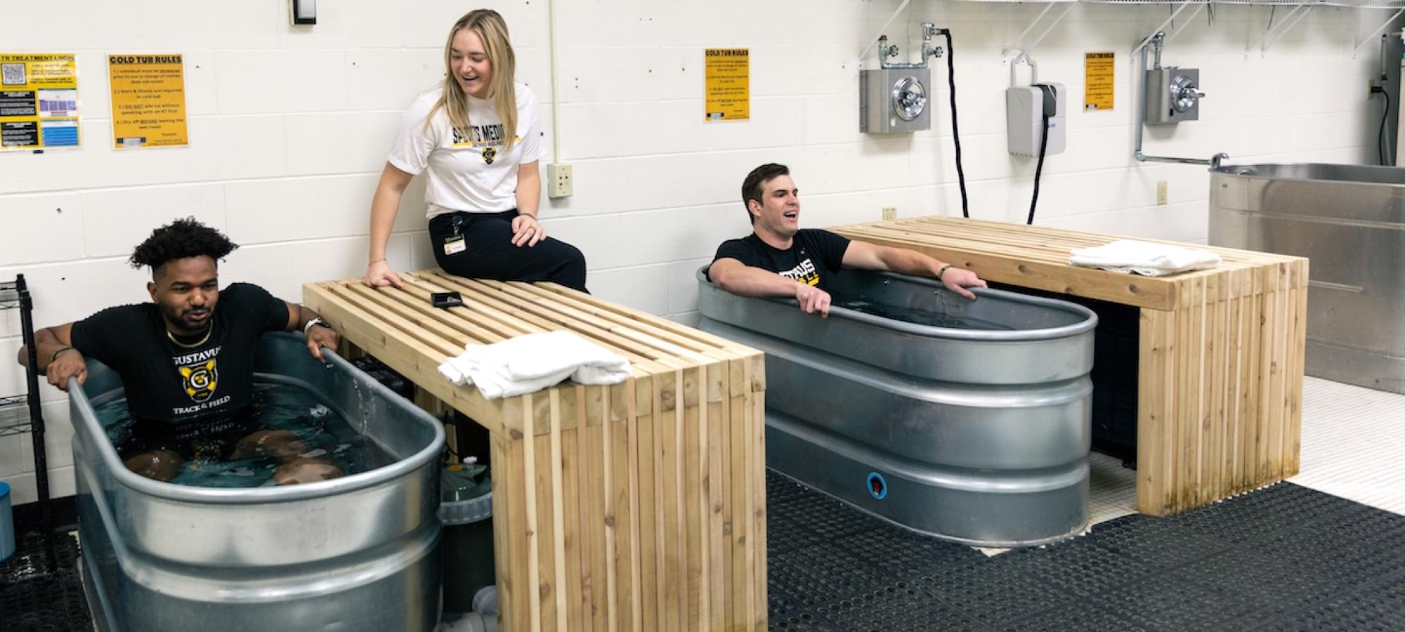 Two athletes in cold submerge tanks are supervised by a trainer. 
