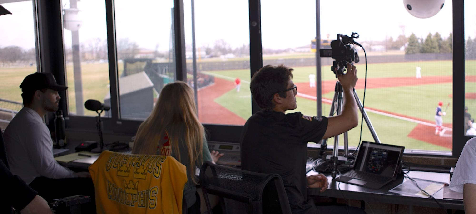 Gustavus Adolphus College sudents in a media booth watching and filming a baseball game. 
