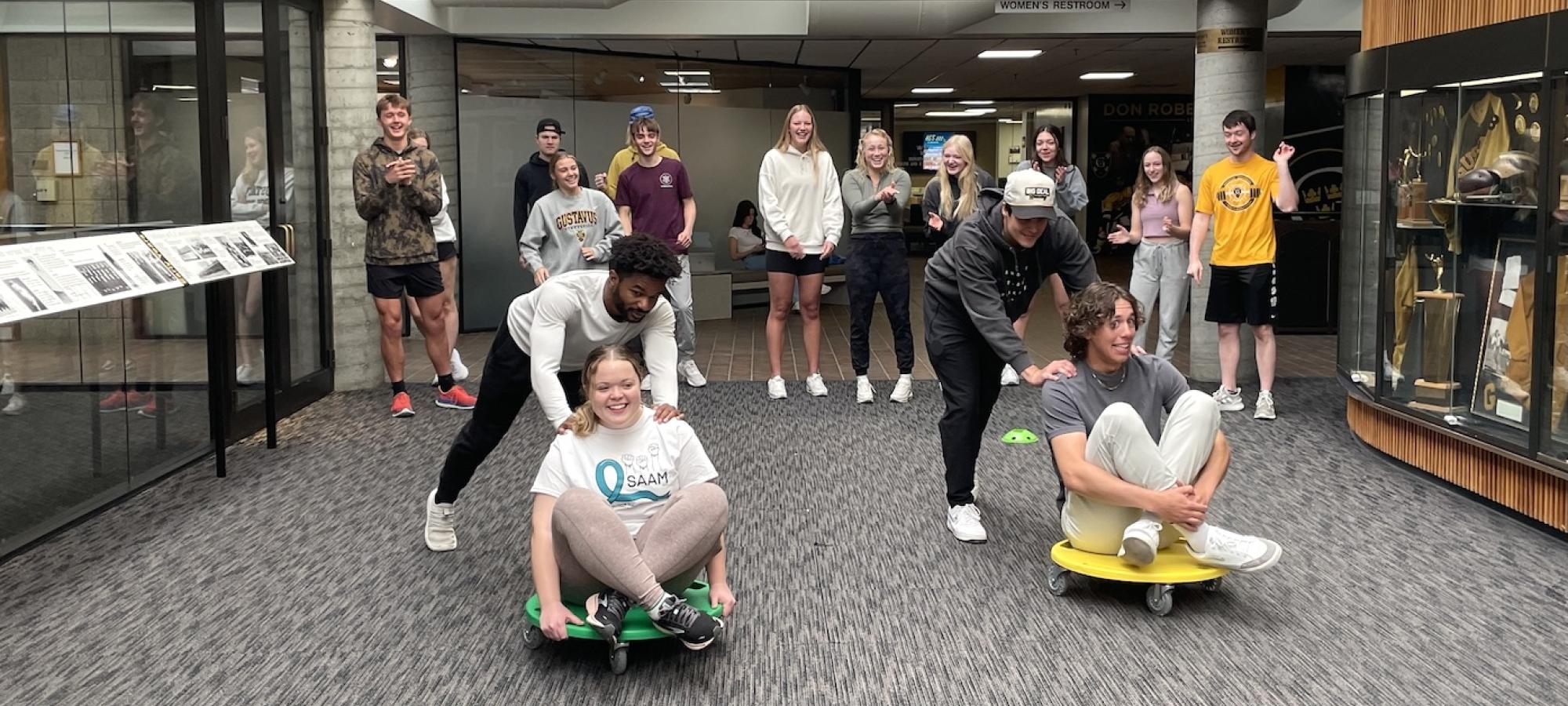 Coaching students push each other on scooters down a hallway.
