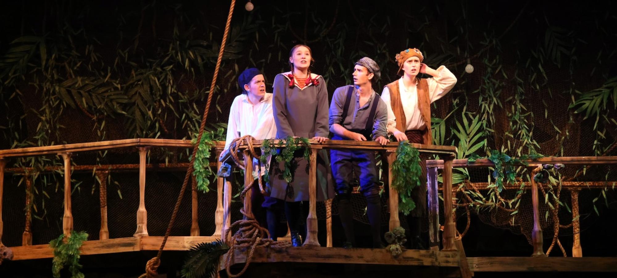 A student in braids sings from a stage bridge, surrounded by a motley crew of young seafarers. 