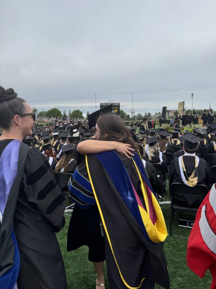 A graduate embraces a faculty member in academic regalia during the Gustavus Adolphus College Commencement ceremony, surrounded by seated graduates in caps and gowns on the football field.