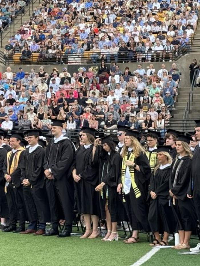A large group of Gustavus Adolphus College graduates in caps and gowns stand on a football field during the Commencement ceremony, facing the crowd-filled bleachers in the background.