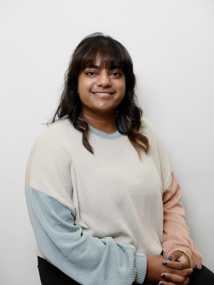 A woman with shoulder-length dark hair and bangs smiles while seated against a plain white background, wearing a color-blocked ribbed sweater with blue, cream, and peach sleeves.
