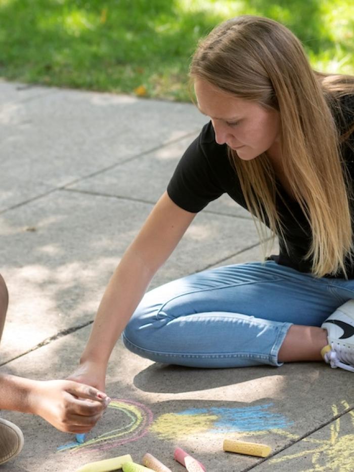 A college student and a child draw with chalk on a sidewalk. 