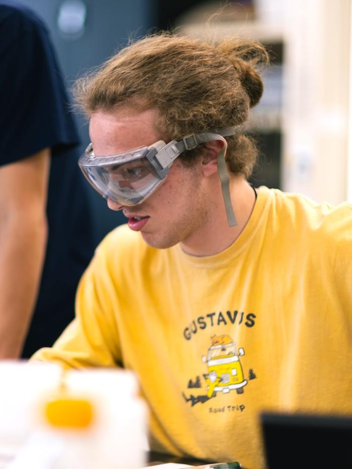 A young man in safety glasses inspects his chemistry work. 