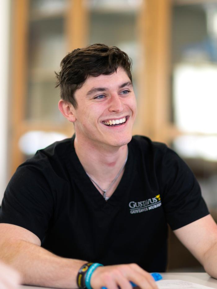 A male Gustavus nursing student in scrubs smiles at something off camera. 