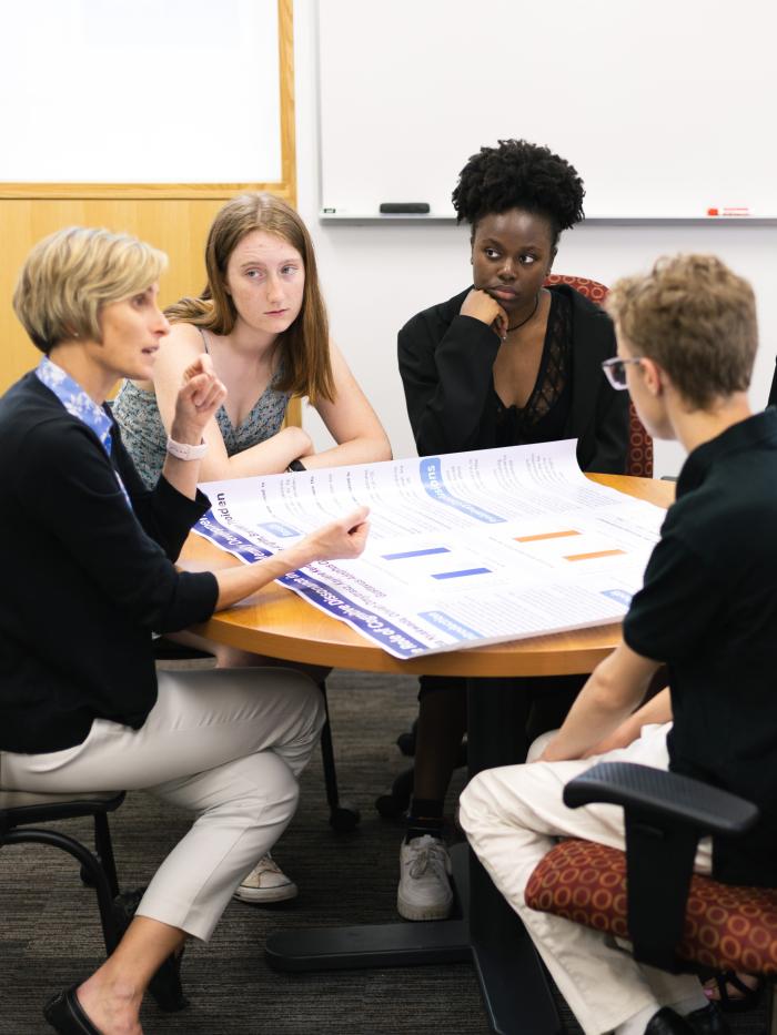 A group of students listens to a professor around a round table. 