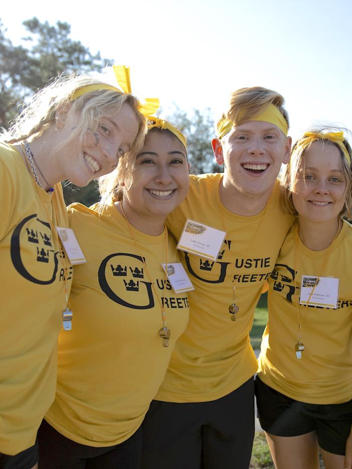 Four students smile at the camera wearing Gustie Greeters shirts. 