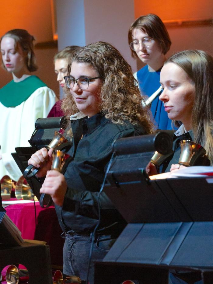 A young woman rings handbells, surrounded by fellow handbell ringers. 